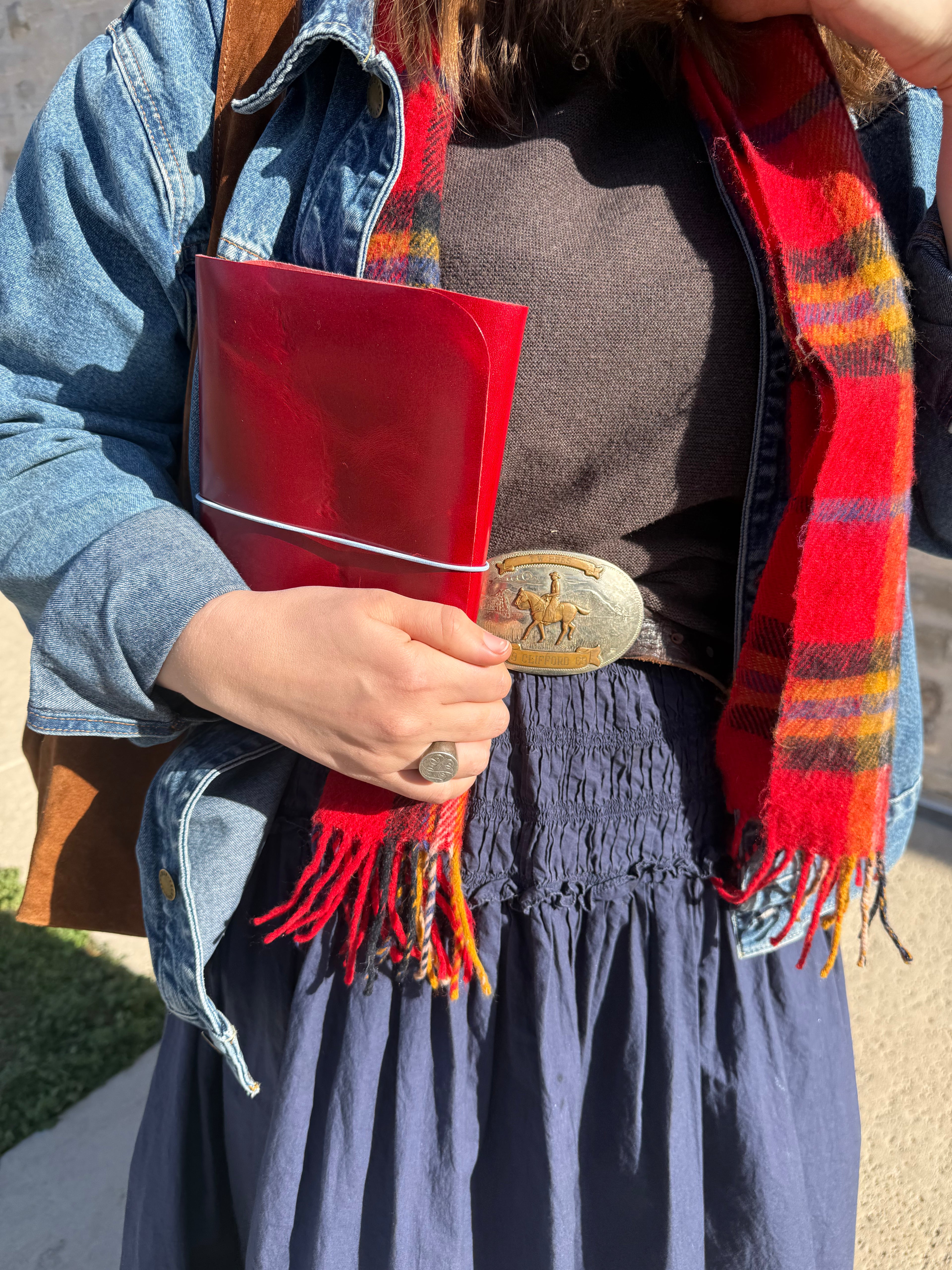 Person holding a red folder with a belt and plaid scarf, wearing a denim jacket.